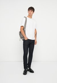 Camiseta blanca de algodón, jeans skinny de denim oscuro, zapatos de cuero negros, mochila gris con correa para el hombro, de pie contra un fondo liso.