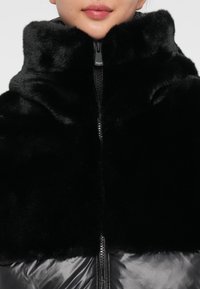 Melnā mākslīgā kažokāda ar augstu apkakli, ar priekšējo rāvējslēdzēju un gludu, spīdīgu audumu apakšējā malā. Mīksta tekstūra visā virsmā.