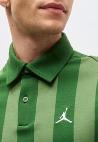 Gros plan d'une personne portant un polo rayé vert avec un logo Jumpman blanc sur la poitrine et une boucle d'oreille à l'oreille gauche.