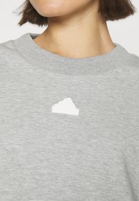 Pilkas džemperis su apvaliu kaklu, pagamintas iš minkšto audinio. Priekyje yra baltas Adidas logotipas.