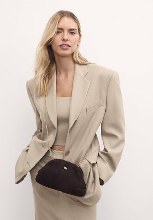 Piccola borsa in suede nera con forma arrotondata, tracolla in catena dorata e dettaglio del logo, indossata sopra un blazer beige e un outfit coordinato.