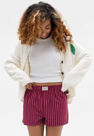 Smilende kvinde med krøllet hår iført hvid kort top, cremefarvet cardigan med grøn bladpatch og shorts med røde striber, der kigger ned på shortsens talje.