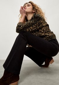 Veste en imprimé léopard, pantalon noir évasé, bottines à talons marron, matériaux texturés, en position accroupie sur un sol gris, fond neutre.