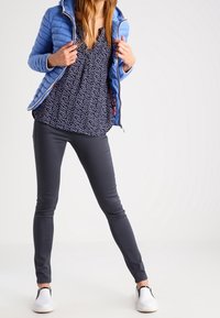 Veste matelassée bleue avec capuche, blouse à pois, pantalon bleu marine et chaussures blanches à enfiler. L'ensemble allie des éléments décontractés avec un look superposé.