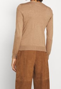 Persona che indossa un maglione beige a maniche lunghe e pantaloni di camoscio marrone, vista da dietro su uno sfondo bianco semplice.