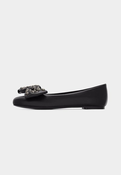 Ballerine plate en cuir noir avec un nœud décoratif orné de clous métalliques sur le bout, vue de profil sur un fond blanc.