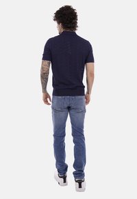 Homme aux cheveux bouclés et tatouages sur le bras gauche, vêtu d'un polo bleu marine, d'un jean bleu et de baskets blanches, debout de dos sur un fond uni.