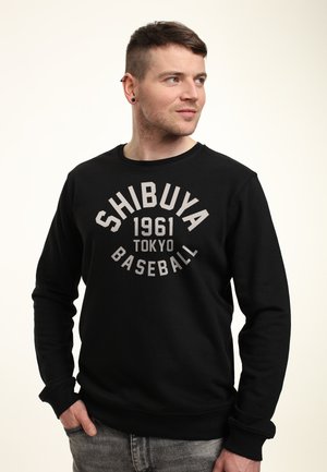 Sweatshirt noir avec un col rond et des manches longues. Texte graphique centré "SHIBUYA 1961 TOKYO BASEBALL" en gris clair. Texture de tissu douce.