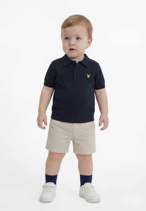 Tout-petit debout portant un polo bleu marine avec un emblème jaune, un short beige, des chaussettes bleu marine et des baskets blanches sur un fond blanc.