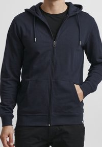 Sweat à capuche zippé bleu marine avec une capuche, des poches avant et des poignets côtelés. Confectionné en tissu doux, présentant une texture lisse et un design minimaliste.