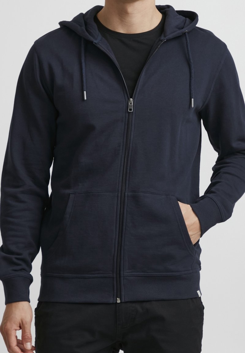 Sweat à capuche zippé bleu marine avec une capuche, des poches avant et des poignets côtelés. Confectionné en tissu doux, présentant une texture lisse et un design minimaliste.