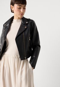 Black leather motorcycle jacket with silver hardware, zippered pockets, and a cropped fit, paired with a cream textured top and skirt.
