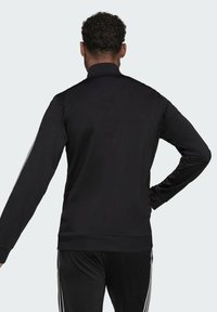 Chaqueta de chándal negra con mangas largas, cuello alzado y rayas plateadas a lo largo de las mangas. Textura de tela suave y diseño ajustado.