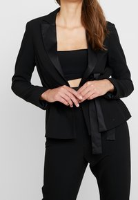 Blazer negro con solapas de satén, diseño ajustado y cintura entallada. Cuenta con un bajo con peplum y se combina con un top corto negro.