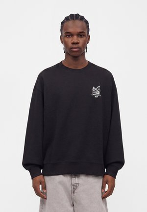 Млад мъж с плитки, облечен в червен суитшърт Carhartt WIP и светлосиви панталони на еднороден бял фон.