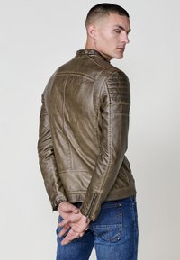 Uomo con giacca di pelle marrone e jeans blu che guarda di lato, con le mani congiunte dietro la schiena, su uno sfondo chiaro e uniforme.