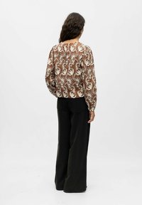 Bruine blouse met bloemenpatroon en lange mouwen, voorzien van crèmekleurige borduursels, gecombineerd met zwarte wijde broek. Achteraanzicht.