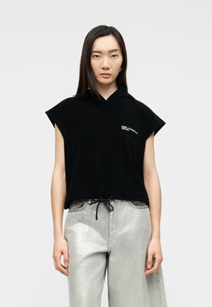 Tânără purtând un hanorac negru fără mâneci cu textul „KARL LAGERFELD JEANS” și pantaloni evazați argintii, stând pe un fundal gri.