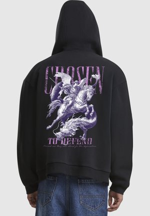 Hoodie noir avec un graphisme au dos représentant un ange sur un cheval affrontant un dragon, avec un texte violet disant "CHOISIE POUR DÉFENDRE". Matière en mélange de coton.