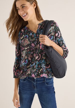 Usměvavá žena v květované blůze a modrých džínách, držící černou polstrovanou kabelku přes rameno na jednobarevném pozadí.