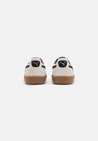 Weiße Wildledersneaker mit schwarzen Akzenten, ausgestattet mit einer beige Gummisohle und einem goldenen Logo auf der Ferse. Die Rückansicht zeigt die glatte Textur und das schlichte Design.