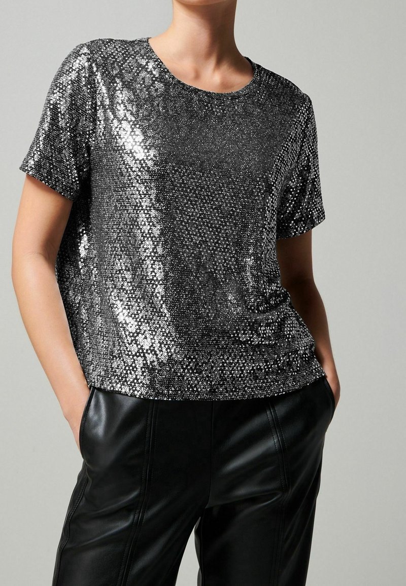 Femme portant un top à manches courtes scintillant argenté en sequins, associé à un pantalon en cuir noir, debout les mains dans les poches.