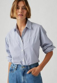 Camicia a righe blu e bianche con colletto alla francese, maniche lunghe arrotolate ai polsi e una morbida texture di cotone. Indossata con jeans a vita alta.