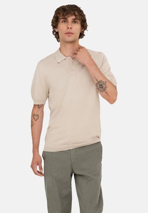 Ung man med lockigt hår, klädd i beige kortärmad pikétröja och grå byxor, visar tatueringar på båda underarmarna mot en vit bakgrund.