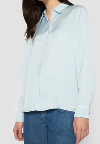 Camicia azzurro chiaro con bottoni realizzata in tessuto morbido. Presenta un colletto a punta, maniche lunghe e un delicato dettaglio della tasca frontale. Abbinata a jeans in denim.