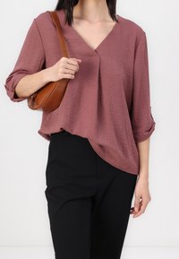 Donker mauve, losvallend V-hals T-shirt met opgerolde mouwen; gecombineerd met zwarte broek en een gestructureerde bruine schoudertas.