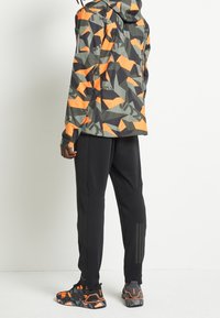 Giacca a fantasia mimetica verde, grigia e arancione; pantaloni sportivi neri; scarpe da ginnastica nere e arancioni con suole testurizzate.