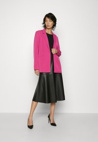 Pink blazer with a tailored fit, worn over a black top and paired with a black leather skirt. Model wears black high heels.