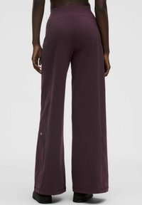 Osoba v širokých tmavofialových nohaviciach s vysokým pásom a čiernych topánkach, stojí s uvoľnenými rukami po stranách na jednofarebnom pozadí.