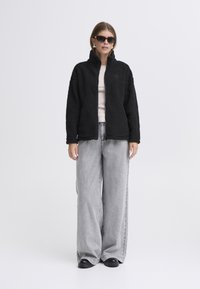 Mustast fliisist jakk kõrge kraega, ees lukuga ja tekstuurse kangaga. Sobitatud hallide laiade pükstega ja mustade jalanõudega.