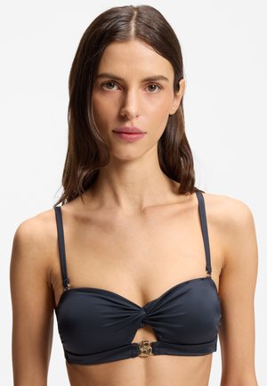 Femme aux longs cheveux bruns portant un haut de bikini noir à fines bretelles et un fermoir doré au centre.