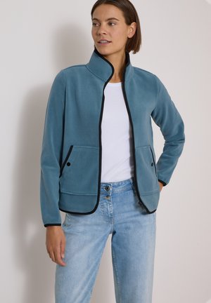 Femme portant une veste polaire bleue zippée avec une bordure noire par-dessus une chemise blanche et un jean bleu clair, posant avec une main sur la hanche.