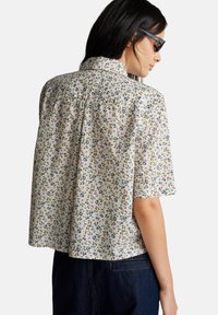 Vrouw die een zonnebril draagt en een kortemouwen, knoopshirt met een klein bloemenpatroon, gecombineerd met donkere spijkerbroeken.