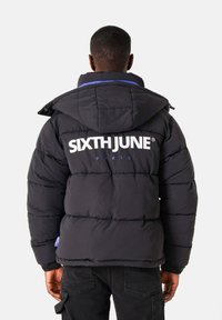 Zwarte pufferjack met capuchon, blauwe accenten en grote witte "SIXTHJUNE" branding op de achterkant. Gestructureerd gewatteerd ontwerp voor isolatie.