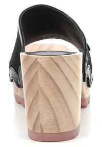Zoccolo in suede nero con tacco in legno, caratterizzato da una finitura liscia, tagli ondulati e borchie in ottone sui lati, su sfondo bianco.