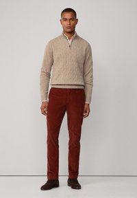 Beige Kabelstrickpullover mit Reißverschluss und Stehkragen, kombiniert mit braunen Cordhosen in Rostfarbe und braunen Dressschuhen.