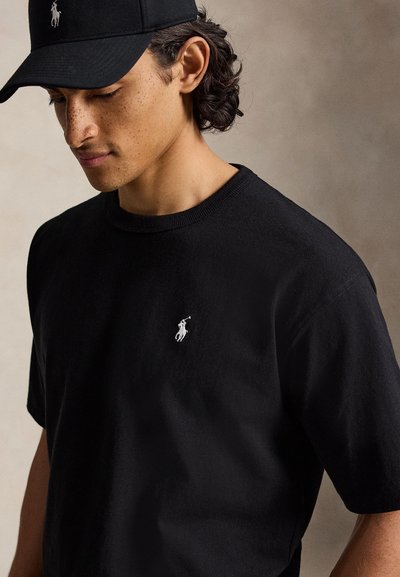 Άνδρας που φοράει μαύρο καπέλο Polo Ralph Lauren και μαύρο T-shirt με λευκό λογότυπο παίκτη πόλο, κοιτάζει προς τα κάτω σε μονόχρωμο φόντο.