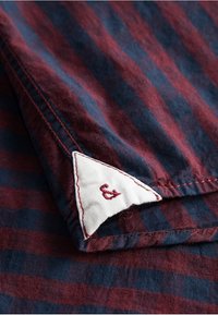 Tela a rayas en tonos marrón y azul marino con una textura sutil. Etiqueta triangular blanca con costura roja que presenta la letra "&" bordada.