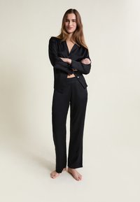 Kobieta stoi boso z założonymi rękami, nosząc czarną, satynową koszulę zapinaną na guziki oraz pasujące luźne spodnie na tle jednolitego jasnego tła.