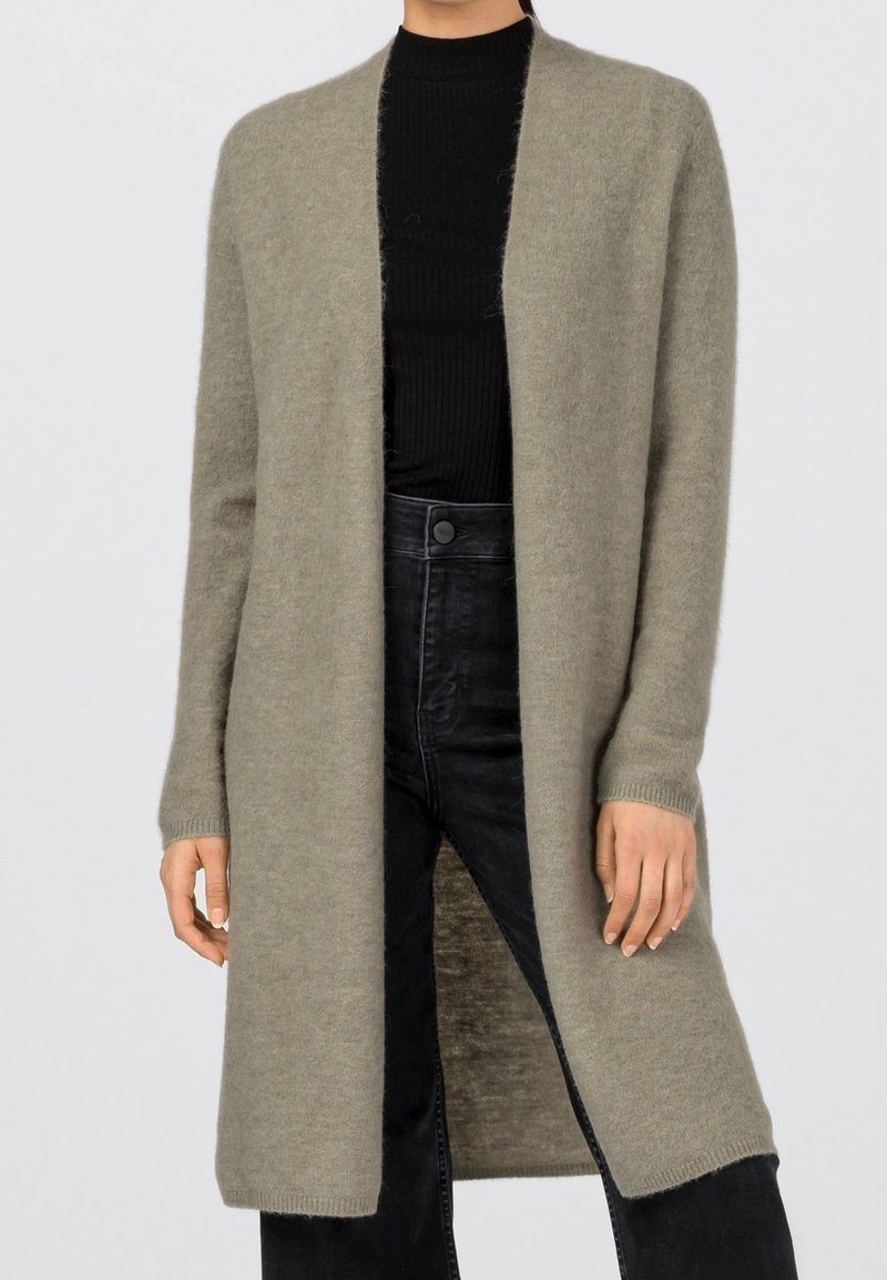 Femme portant un long cardigan gris ouvert sur un pull à col roulé noir côtelé et un jean noir taille haute.