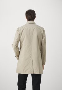 Beige vettcoats vedenkestävä trench-coat, jossa on suora leikkaus, näkyvät ompeleet ja kaksi etuleikkausta, päällä mustat housut.