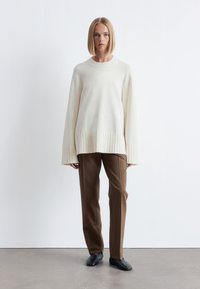 Übergroßer cremefarbener Pullover mit geripptem Saum und Bündchen, kombiniert mit enganliegenden braunen Hosen und schwarzen Schuhen vor einem neutralen Hintergrund.