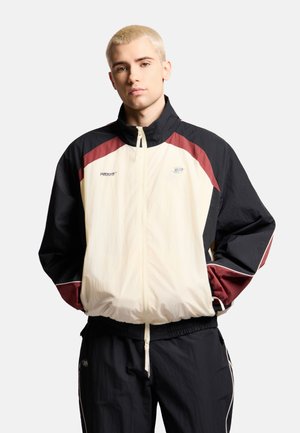 Młody mężczyzna z krótkimi blond włosami, ubrany w czarną, kremową i bordową sportową kurtkę na zamek z rękami w kieszeniach.