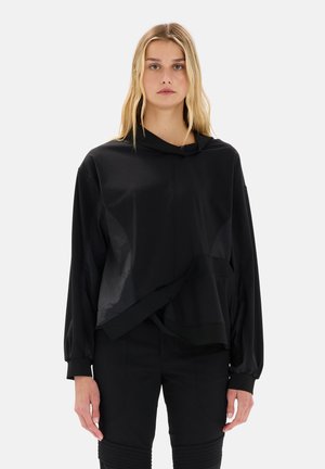 Donna bionda che indossa una maglia nera a maniche lunghe, ampia, con orlo asimmetrico e pantaloni neri, in piedi contro uno sfondo chiaro e uniforme.