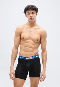 Νεαρός άνδρας με σγουρά μαλλιά, φορώντας μαύρα μποξεράκια Nike με μπλε ζώνη μέσης, στέκε ται μπροστά σε απλό ανοιχτό φόντο.