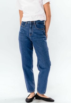 Personne portant un jean en denim bleu taille haute, un t-shirt blanc à manches courtes et des ballerines noires, debout devant un fond clair uni.